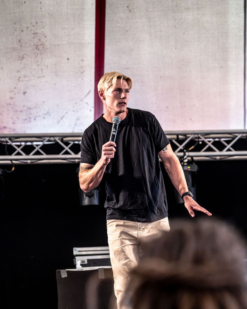 Blond man in black t-shirt and khaki pants speaks into microphone on stage at outdoor event.
