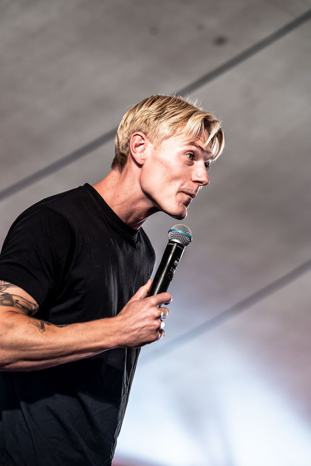Blonde male performer in black t-shirt holding microphone on stage, showing tattoo on arm.