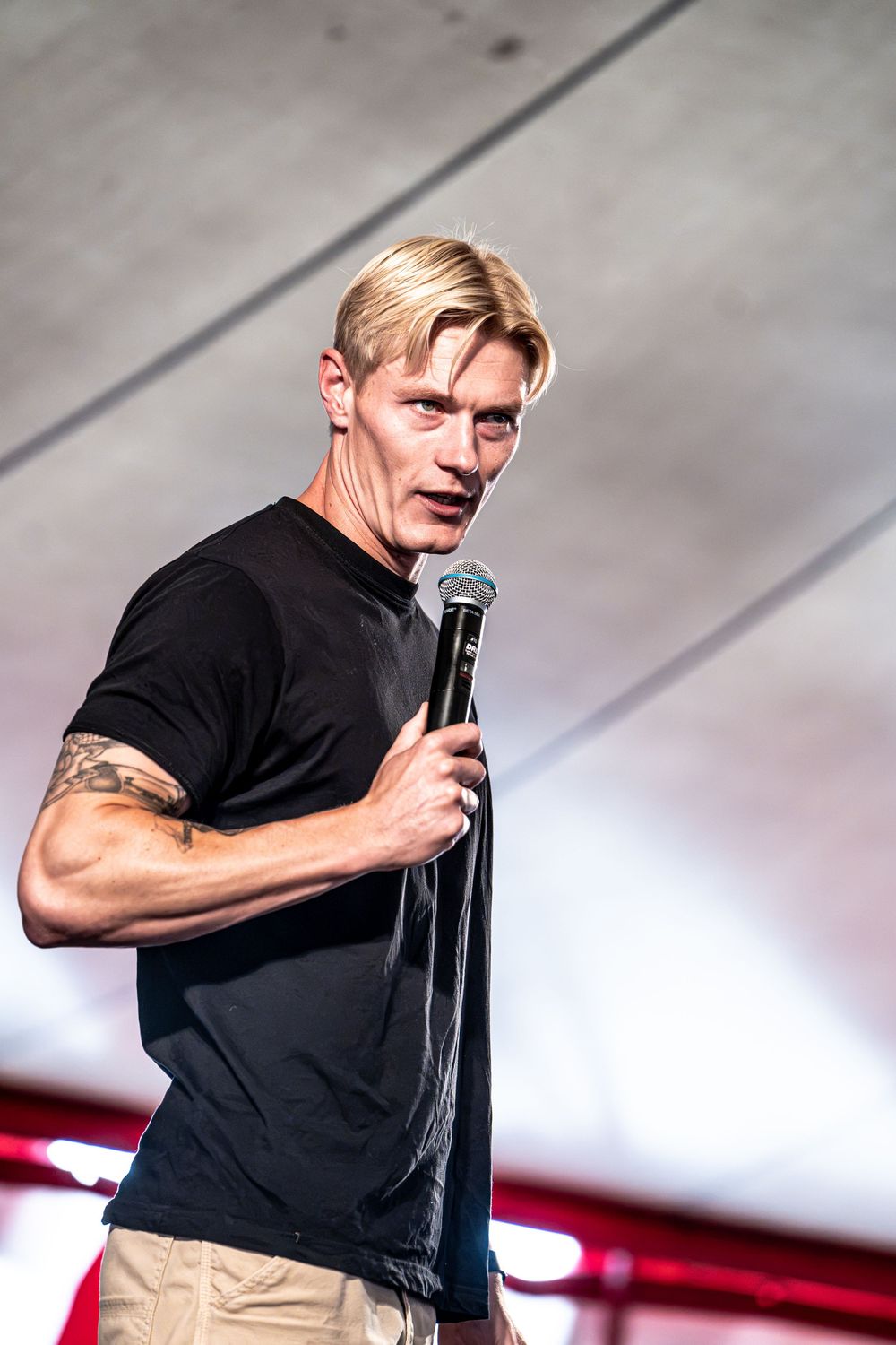 Blonde tattooed male speaker holding microphone on stage in black t-shirt.