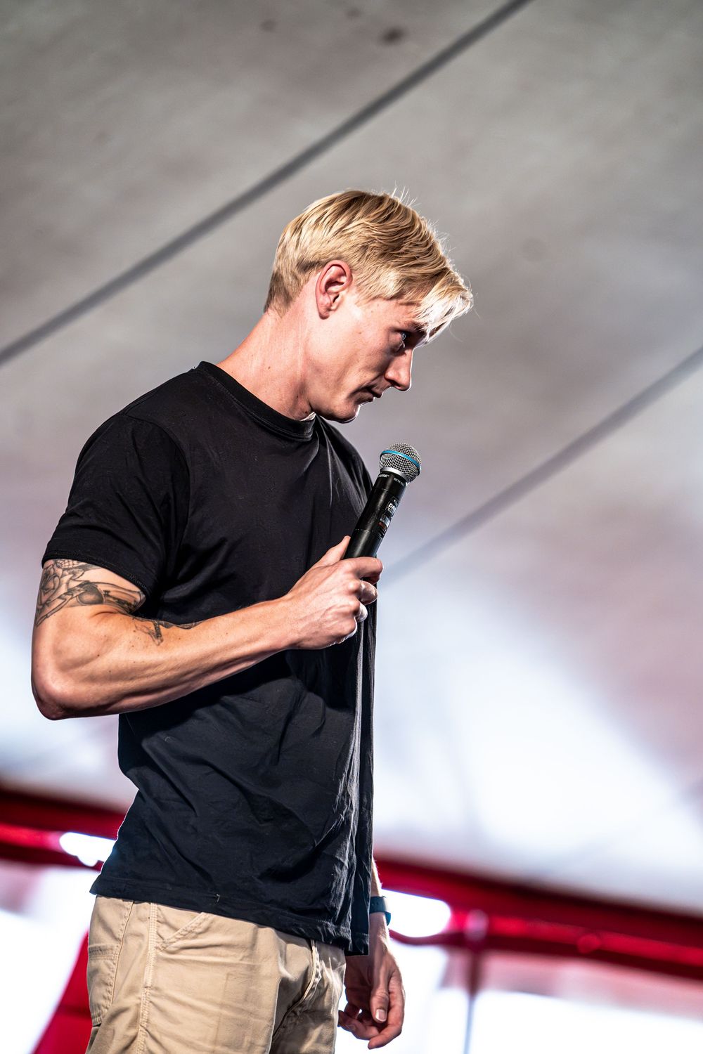 Blond male performer in black t-shirt holding microphone on stage looking down.