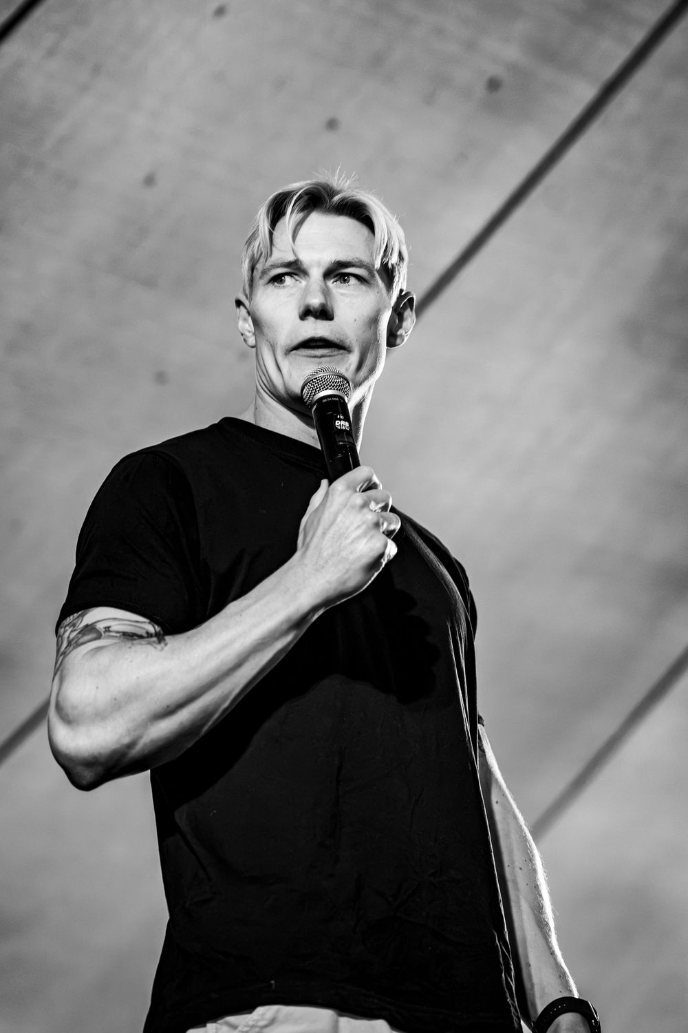 Black and white portrait of blonde tattooed male performer holding microphone on stage.