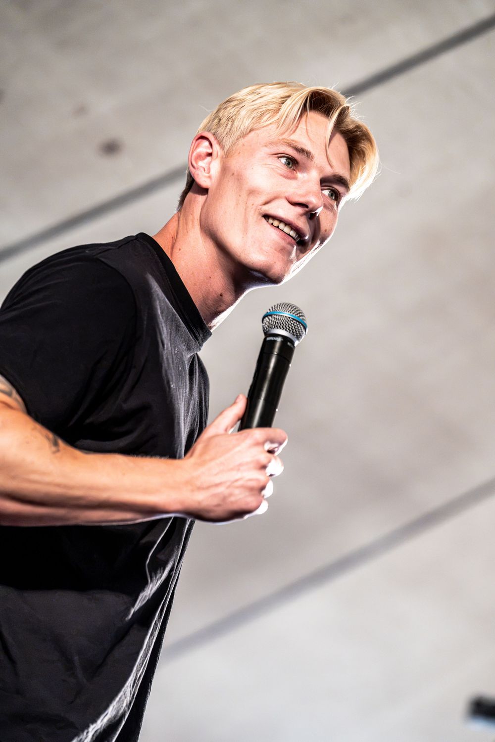 Smiling blond male performer holding microphone on stage wearing black t-shirt.