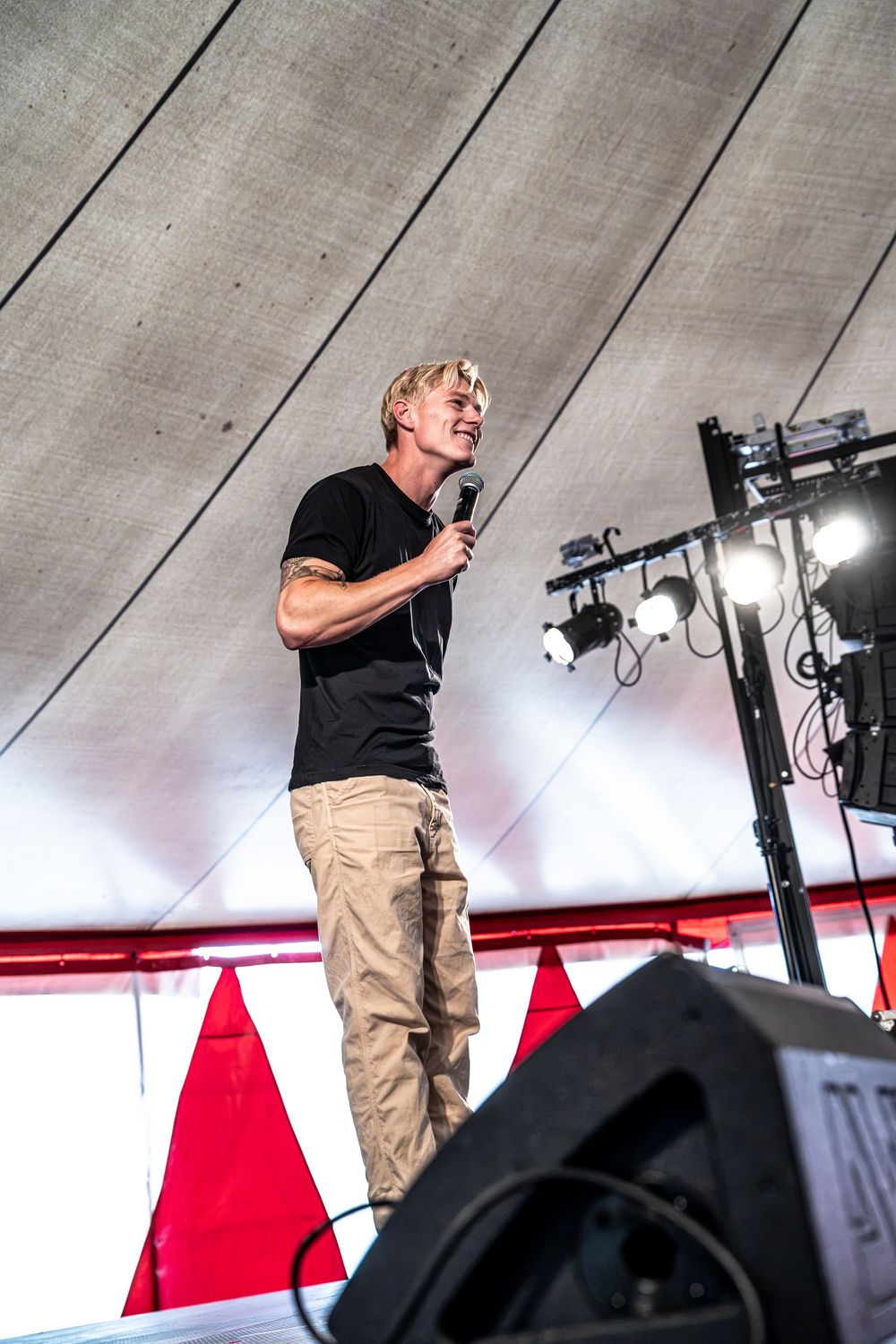Tattooed blonde male performer singing on stage under a tent wearing black shirt and khaki pants at outdoor festival.