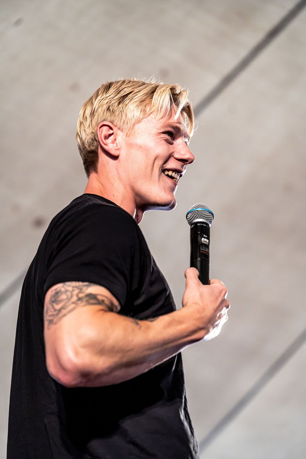 Smiling blond male performer holding microphone on stage during outdoor concert performance.