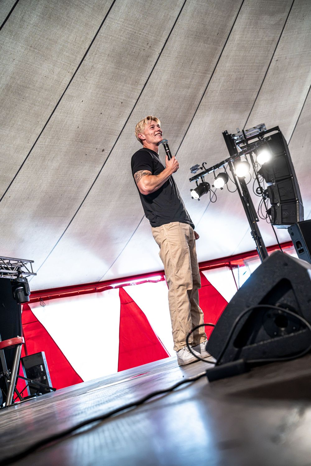 Blond male artist performing on stage with microphone, wearing black shirt and khaki pants, with red and white backdrop.