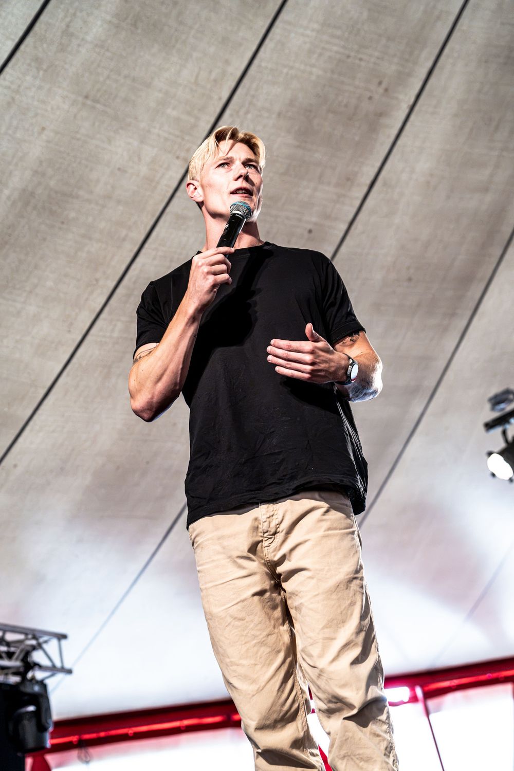 Blonde man in black t-shirt and khaki pants speaks into microphone on stage.