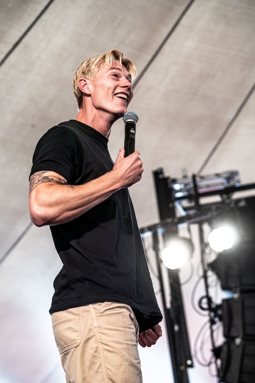 Laughing blond male performer holding microphone on stage wearing black t-shirt and khaki pants.