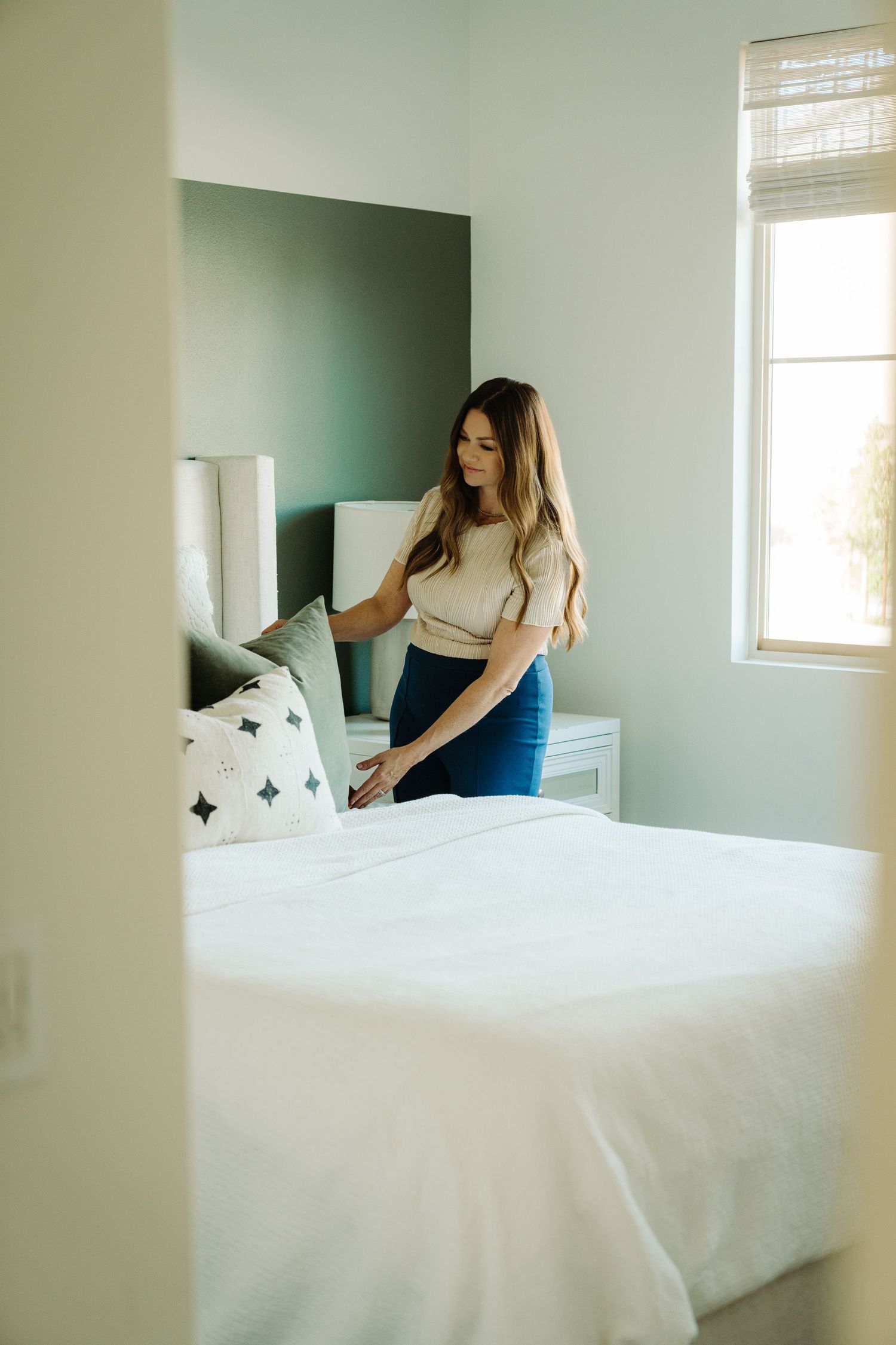 Woman making a bed in a modern bedroom with green accent wall, white bedding, and decorative pillows near a bright window.