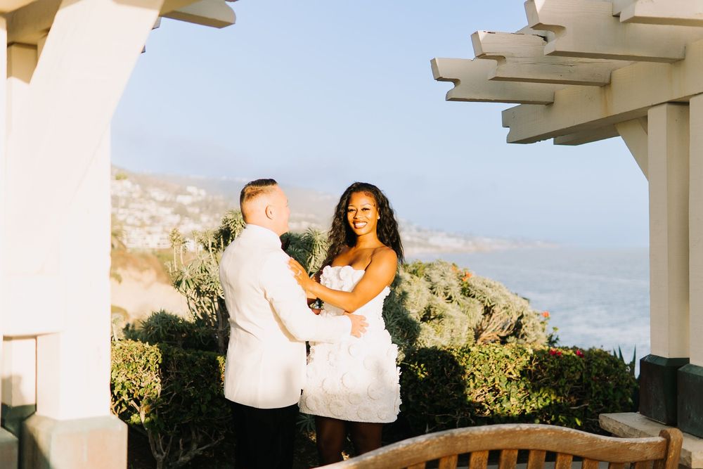 Couple holds hands during outdoor wedding ceremony with ocean and coastal views in background on sunny day.