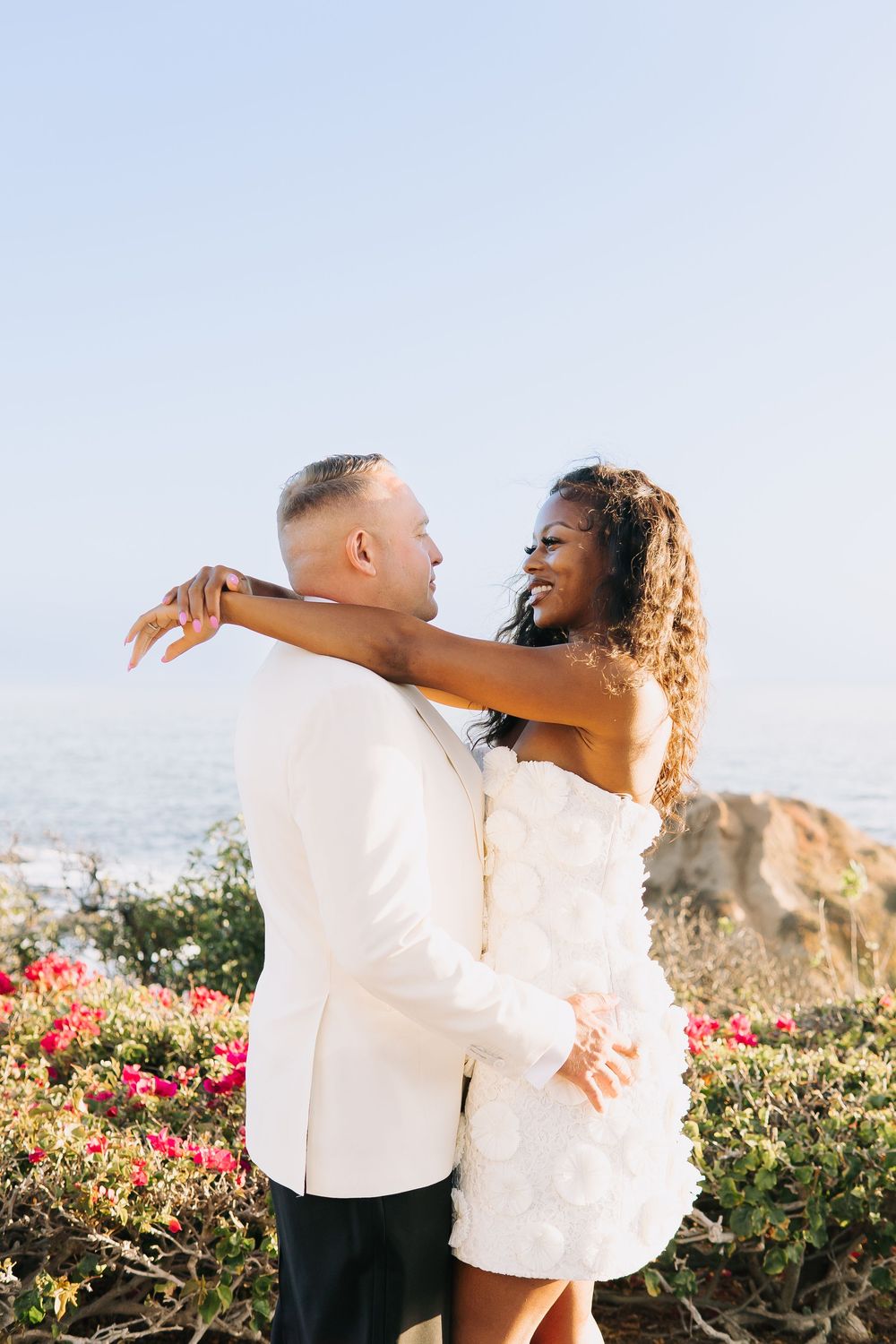 A couple in white attire embraces outdoors near the ocean, surrounded by colorful flowers under a clear sky.