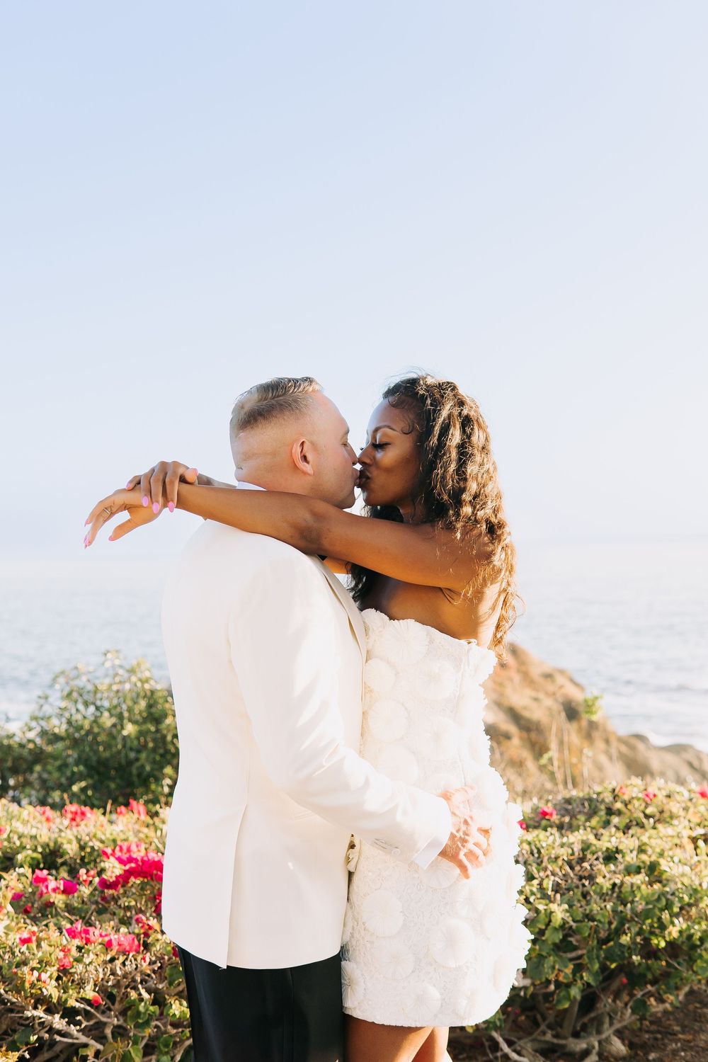 Couple in white wedding attire sharing a kiss by the ocean with colorful flowers in the background.