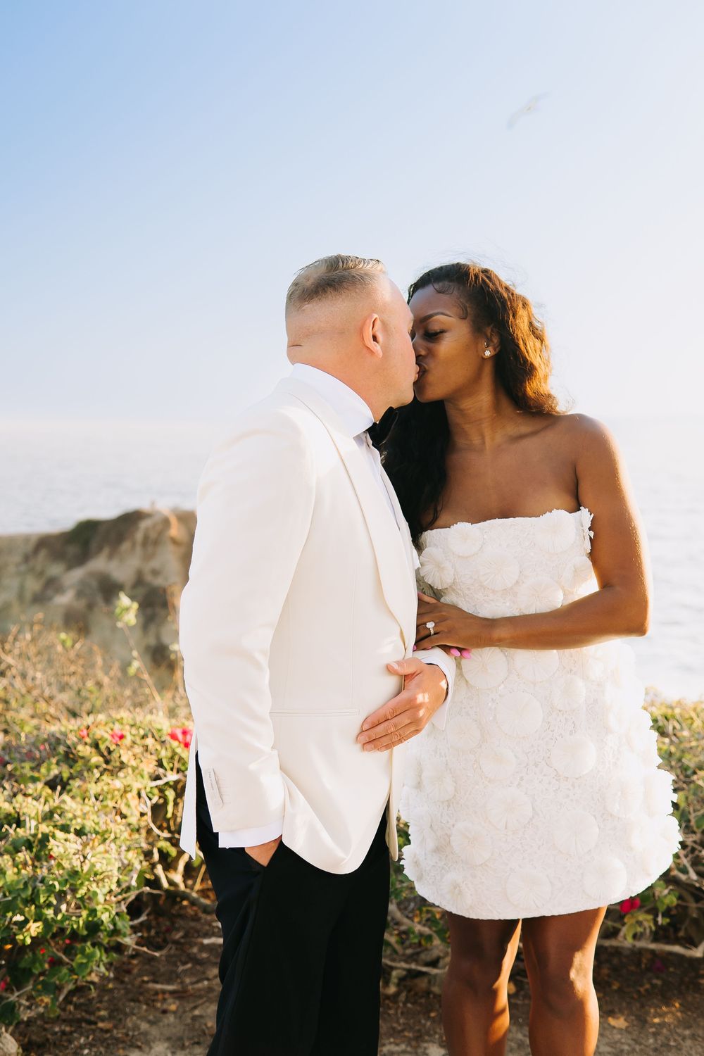 Interracial couple kissing in white wedding outfits on a cliffside overlooking the ocean.
