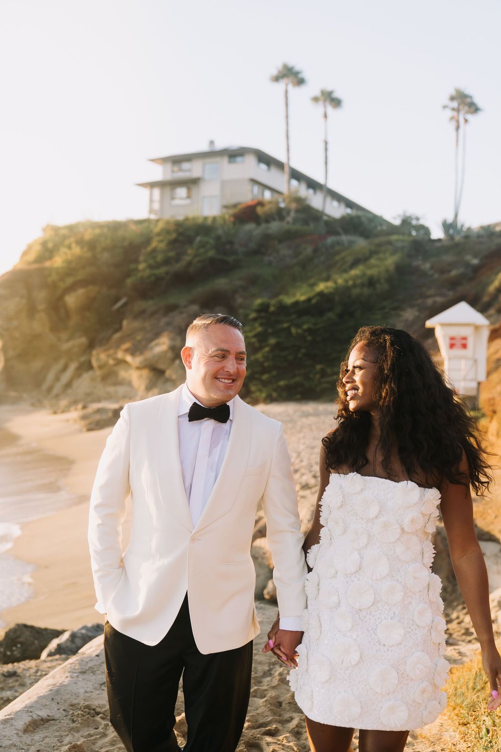 Interracial couple in wedding attire holding hands on a sunny beach with palm trees and cliffside home.