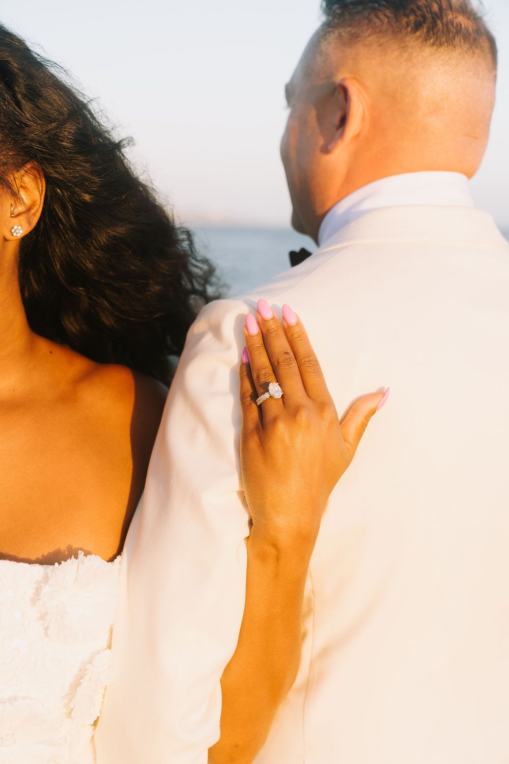 Bride places hand on groom's back showcasing elegant engagement ring, pink nails, at sunset beach wedding.
