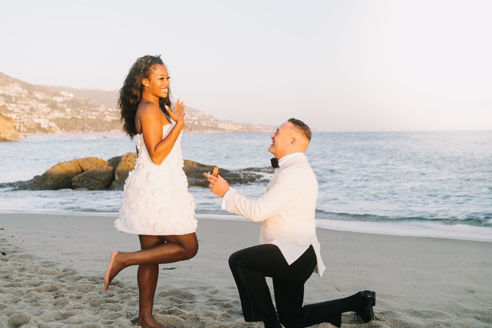 Man proposes to woman on one knee at a scenic beach during golden hour, both dressed in white outfits.