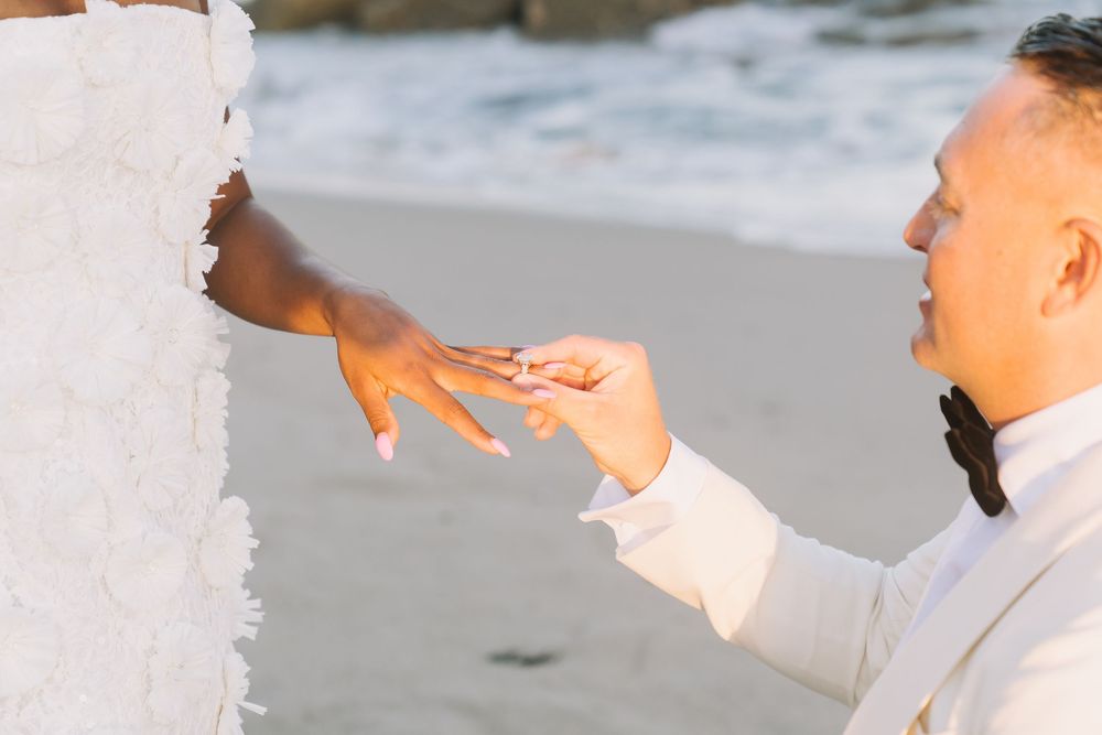 Groom placing a wedding ring on brides hand during a romantic beach ceremony at sunset.