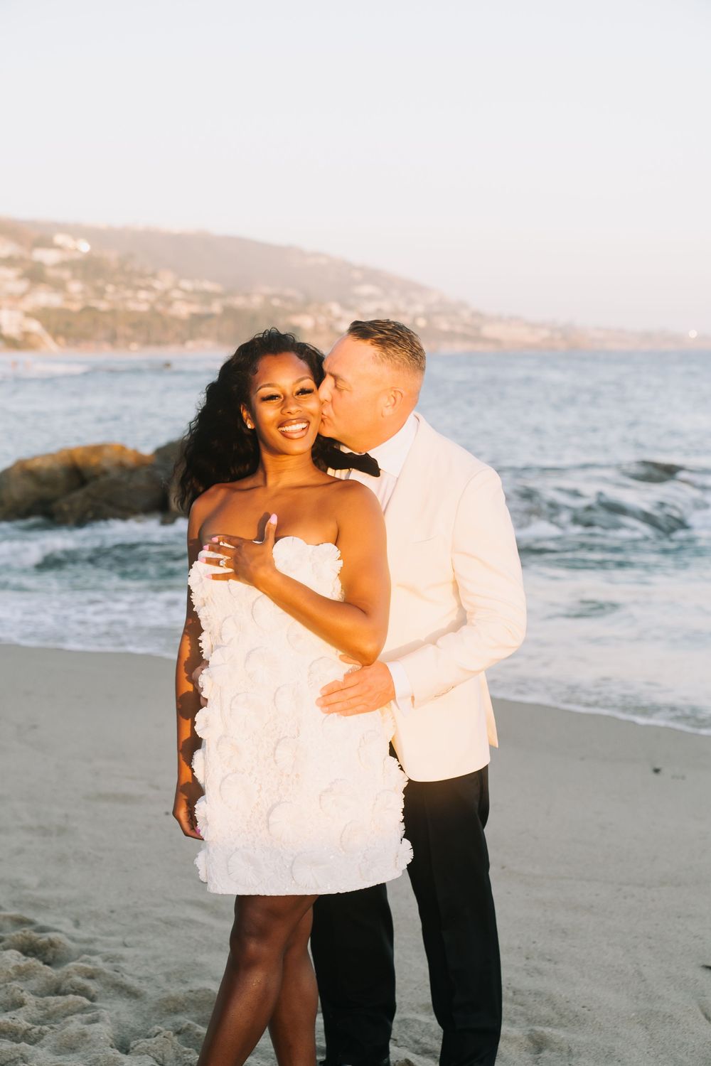 Couple in wedding attire embracing on a sandy beach at golden hour with ocean waves and hills behind them.