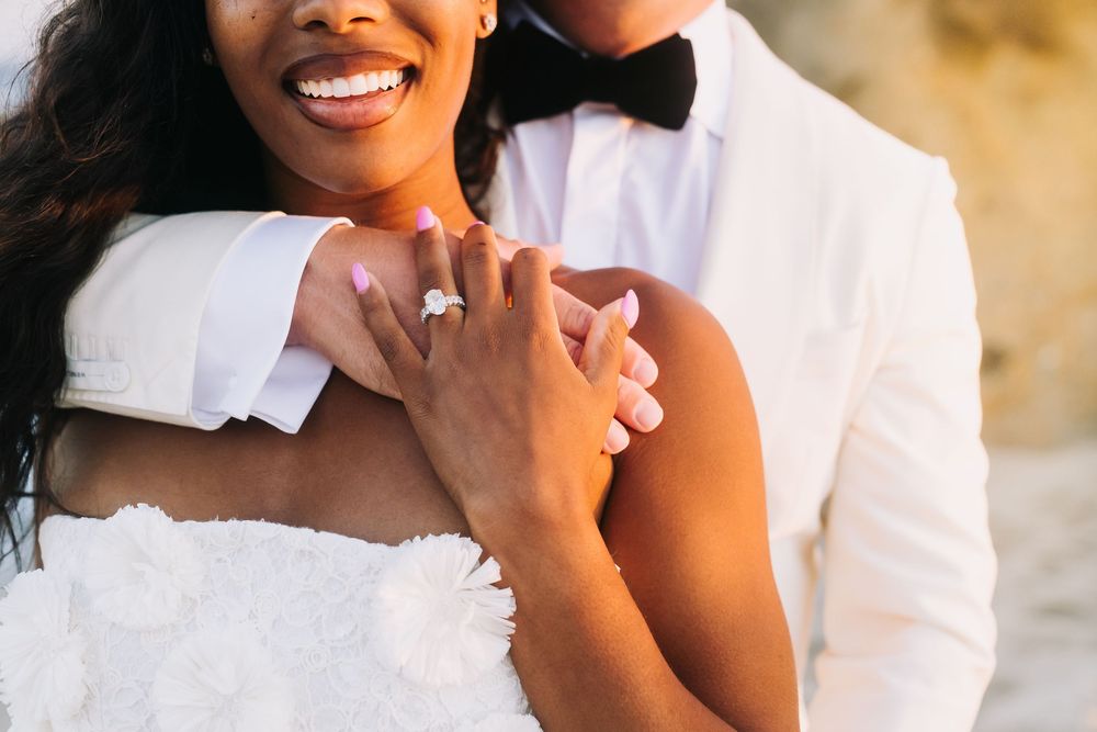 Close-up of bride showing diamond engagement ring while groom in white tuxedo embraces her shoulders.