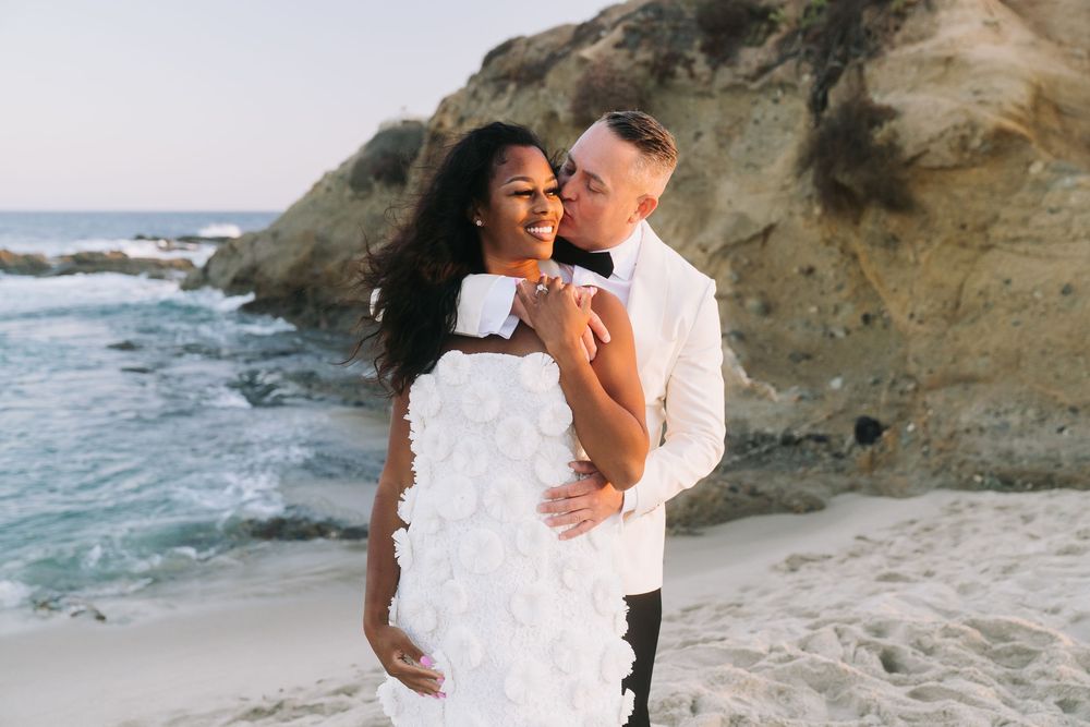 Couple embracing on a beach, bride in white floral dress, groom in white suit, rocky cliffs and ocean waves behind them.