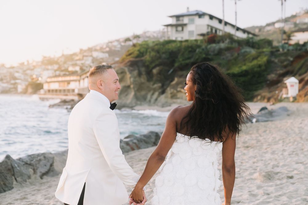 Bride and groom holding hands on a sandy beach at sunset, with rocky cliffs and coastal homes in the background.