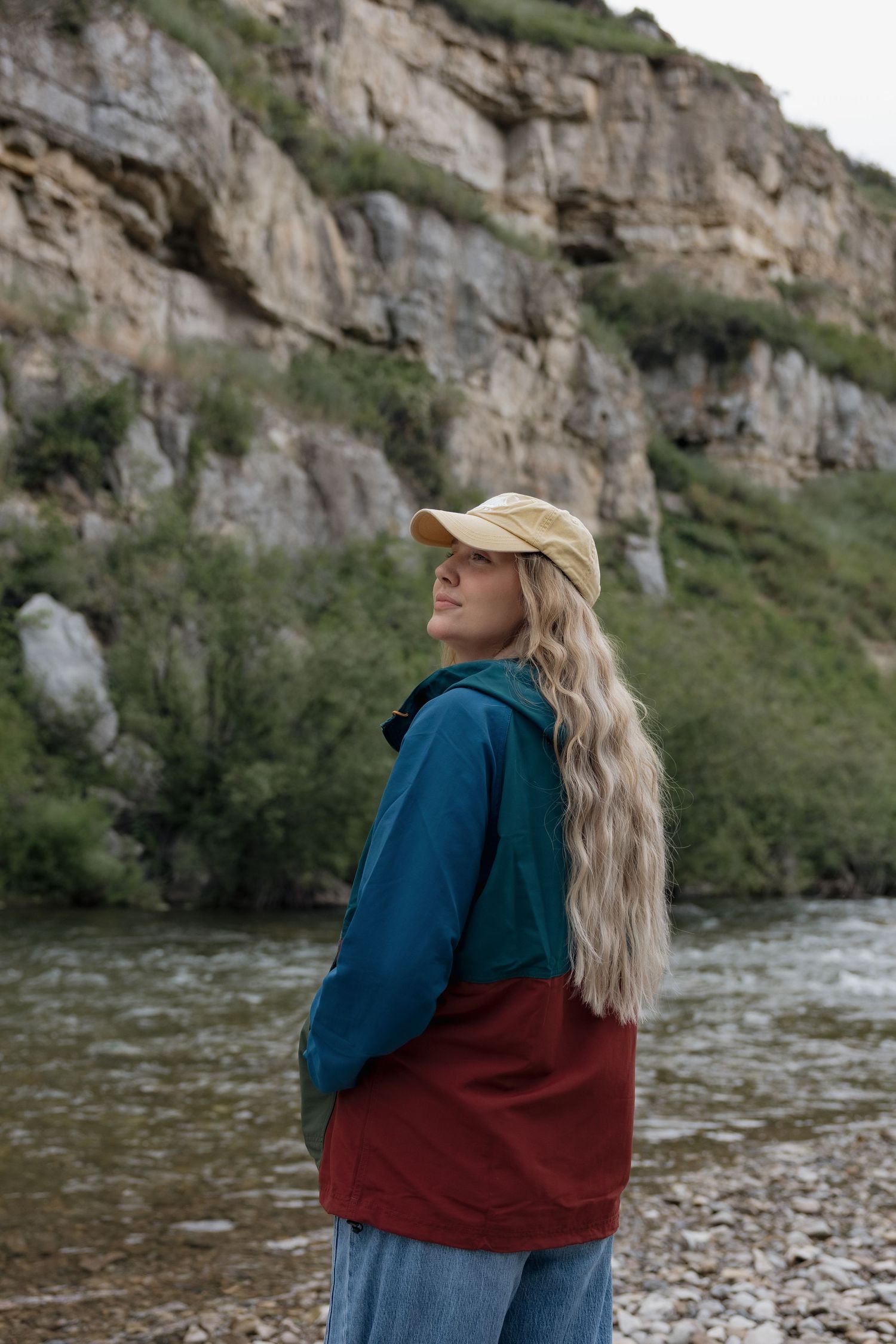Smiling woman with long blonde hair holds a camera outdoors near a rocky riverbank with green hills behind her.