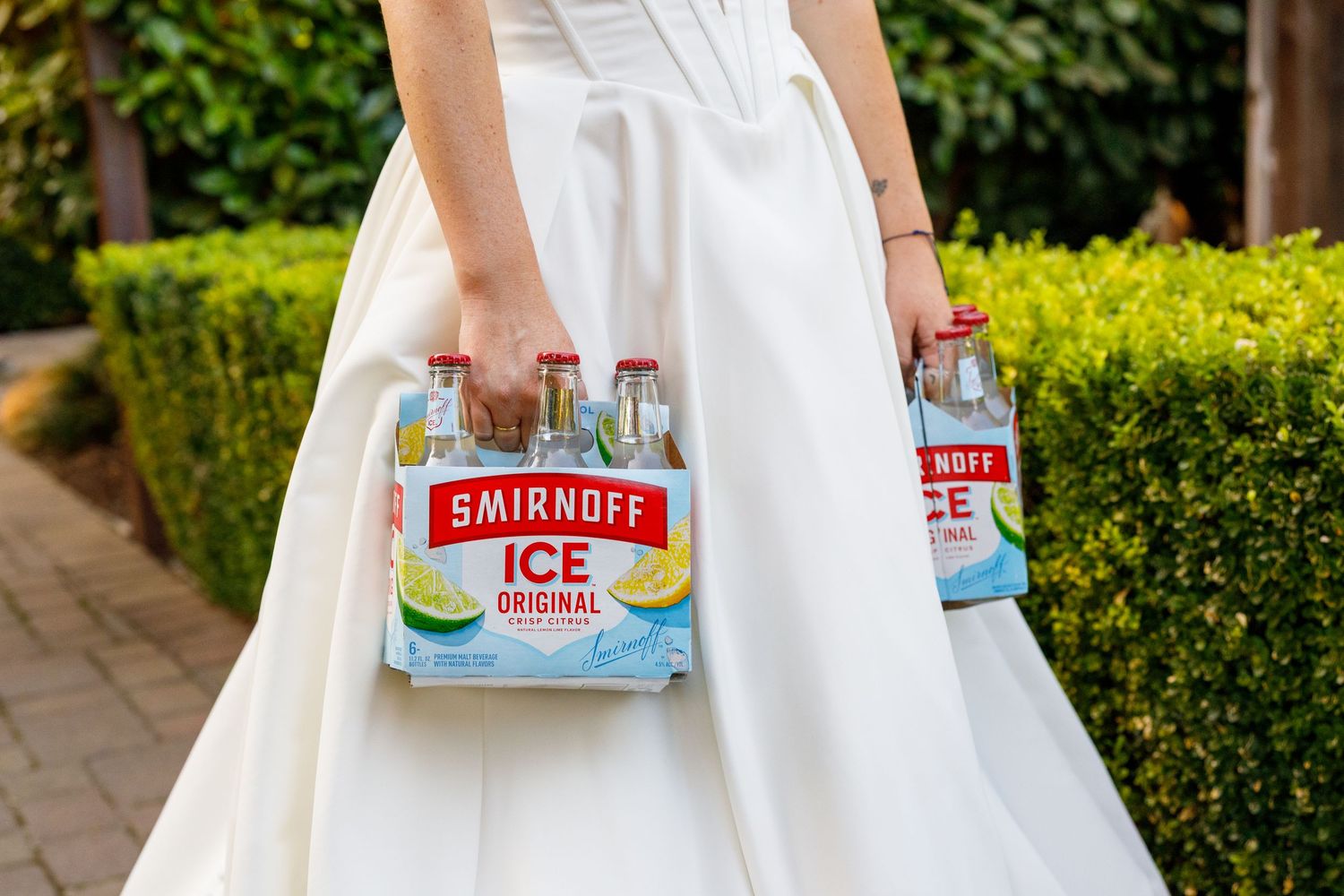 Bride in white gown holding two six-packs of Smirnoff Ice Original bottles outdoors.