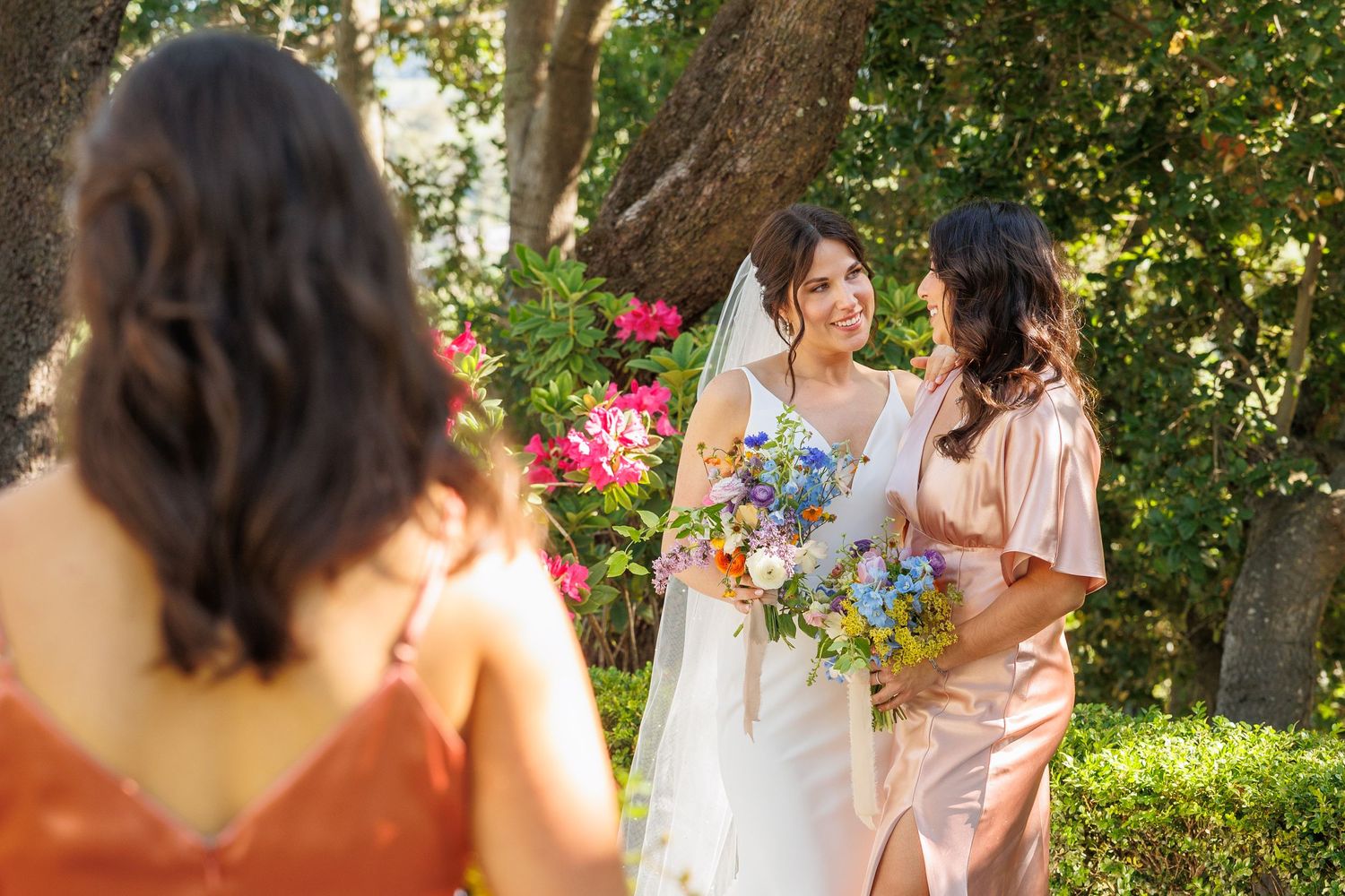 Bride in white gown and veil holds blue bouquet with bridesmaid in pink dress outdoors near colorful flowers.