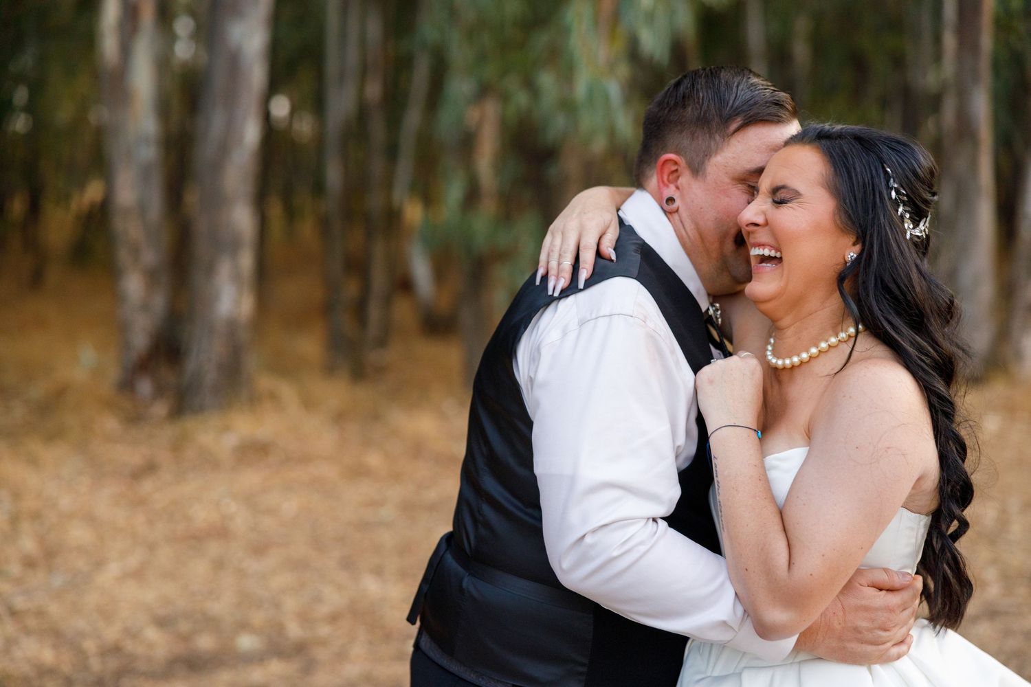 Bride and groom embracing and laughing joyfully in a forest setting, bride wearing pearl necklace and white dress.