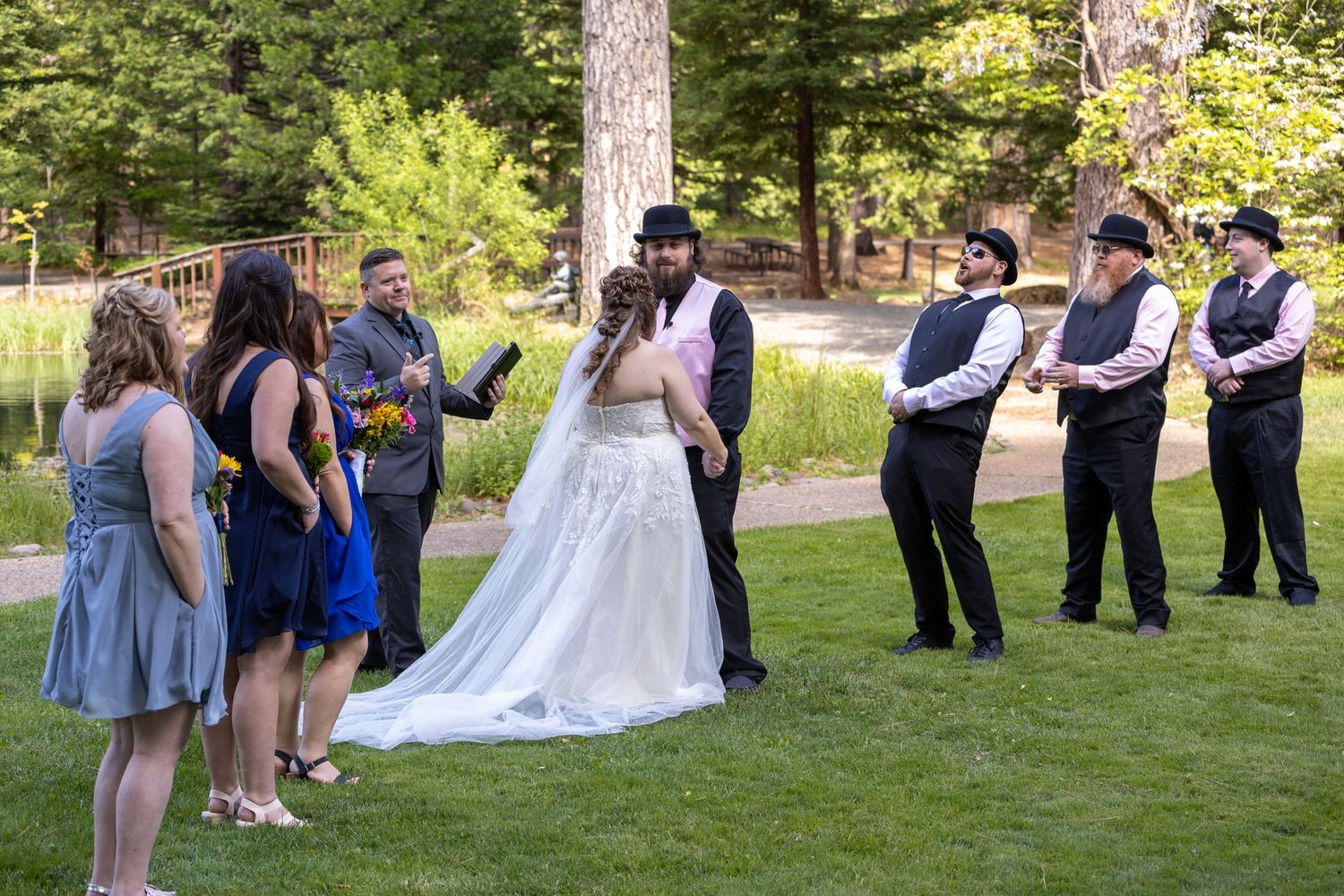 Wedding ceremony outdoors with bride, groom, and wedding party on green lawn among trees.