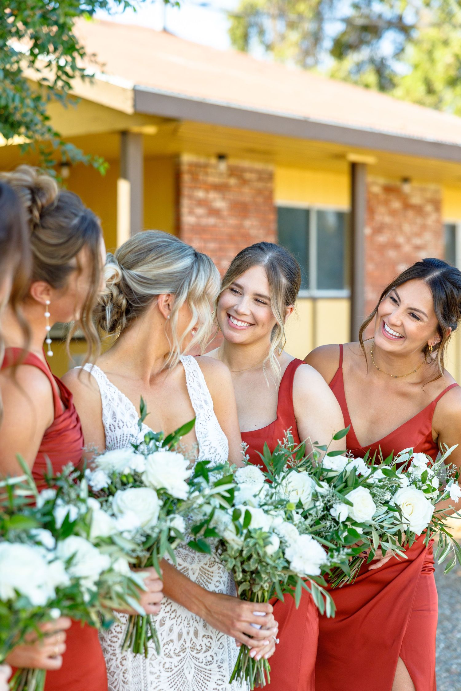 Bride in white lace gown laughing with bridesmaids in red dresses holding white floral bouquets outdoors.