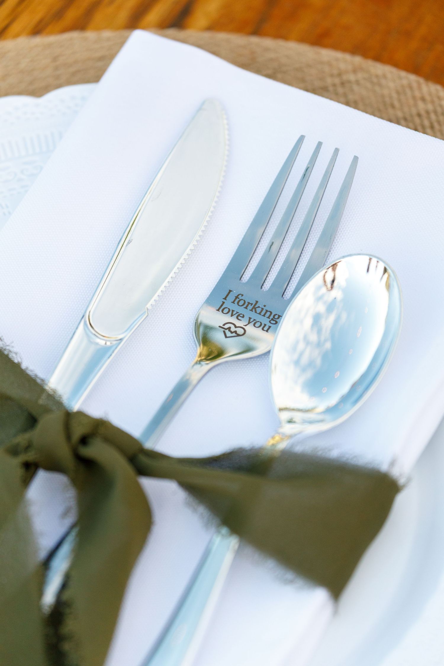 Elegant silver cutlery set with engraved fork reading 'I forking love you' tied with olive green ribbon on white napkin.