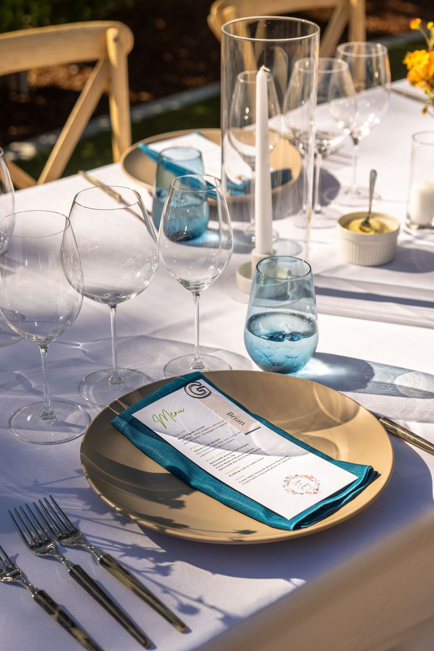 Elegant outdoor dining table set with gold charger plates, teal accents, wine glasses, and a menu card on white linen.