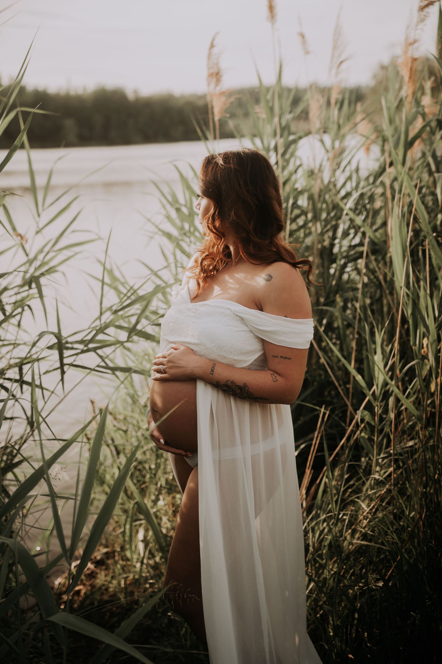 Pregnant woman in white off-shoulder gown standing among tall reeds by a lake during golden hour.