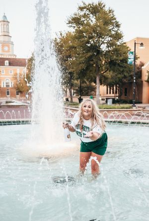 Joyful blonde girl in UNT shirt and green shorts splashing in a campus fountain at golden hour.