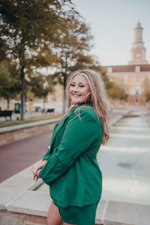 Smiling blonde woman in a green blazer and skirt posing near a fountain on a college campus.