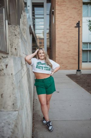 Smiling blonde girl in University of North Texas crop top and green shorts leaning against a brick wall.