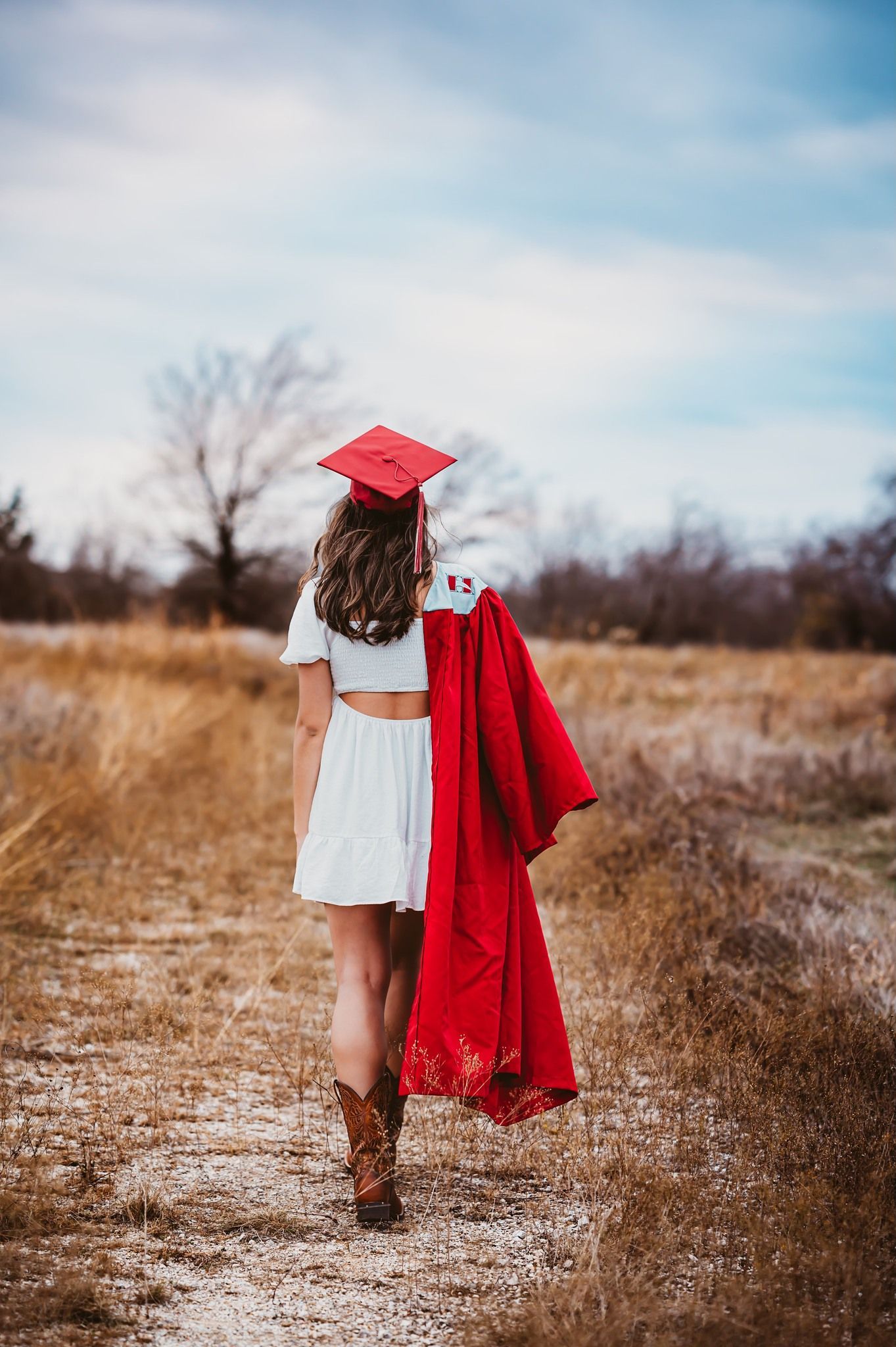 High school senior girl in red graduation cap and gown walking down a rural dirt path.
