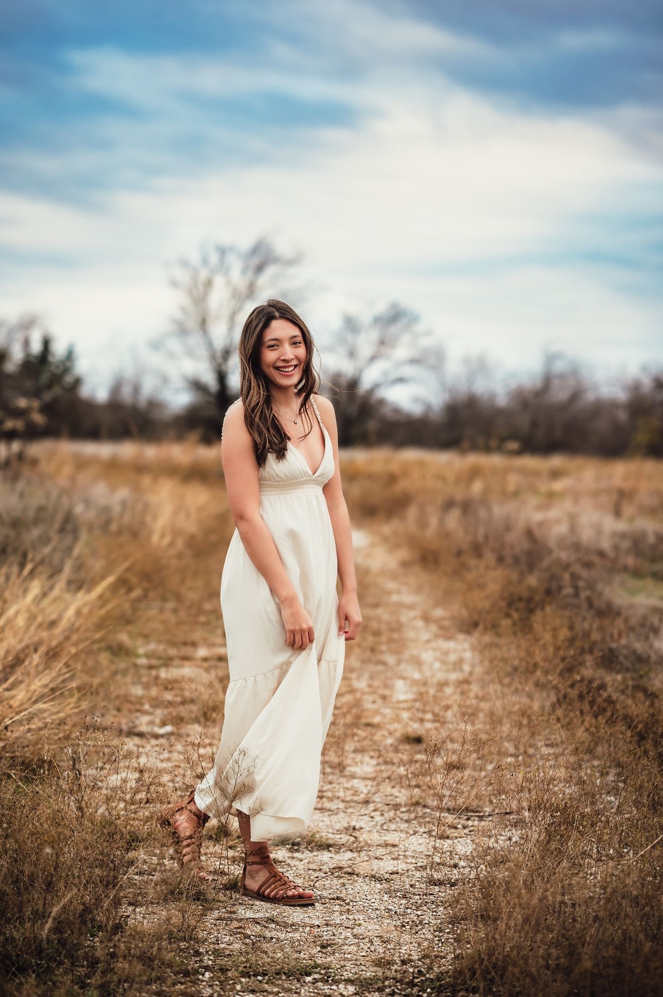 Young woman in a flowing white maxi dress smiling on a rustic dirt path in a field.