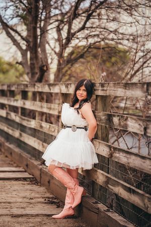 Teen girl in white dress and pink cowboy boots leaning against a wooden bridge railing.