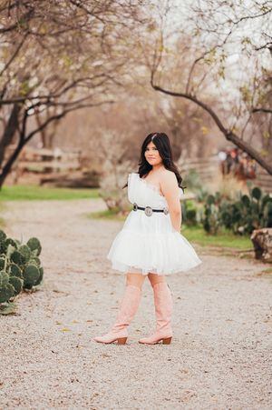 Teen girl in white dress with pink cowboy boots posing on a gravel path near cacti.