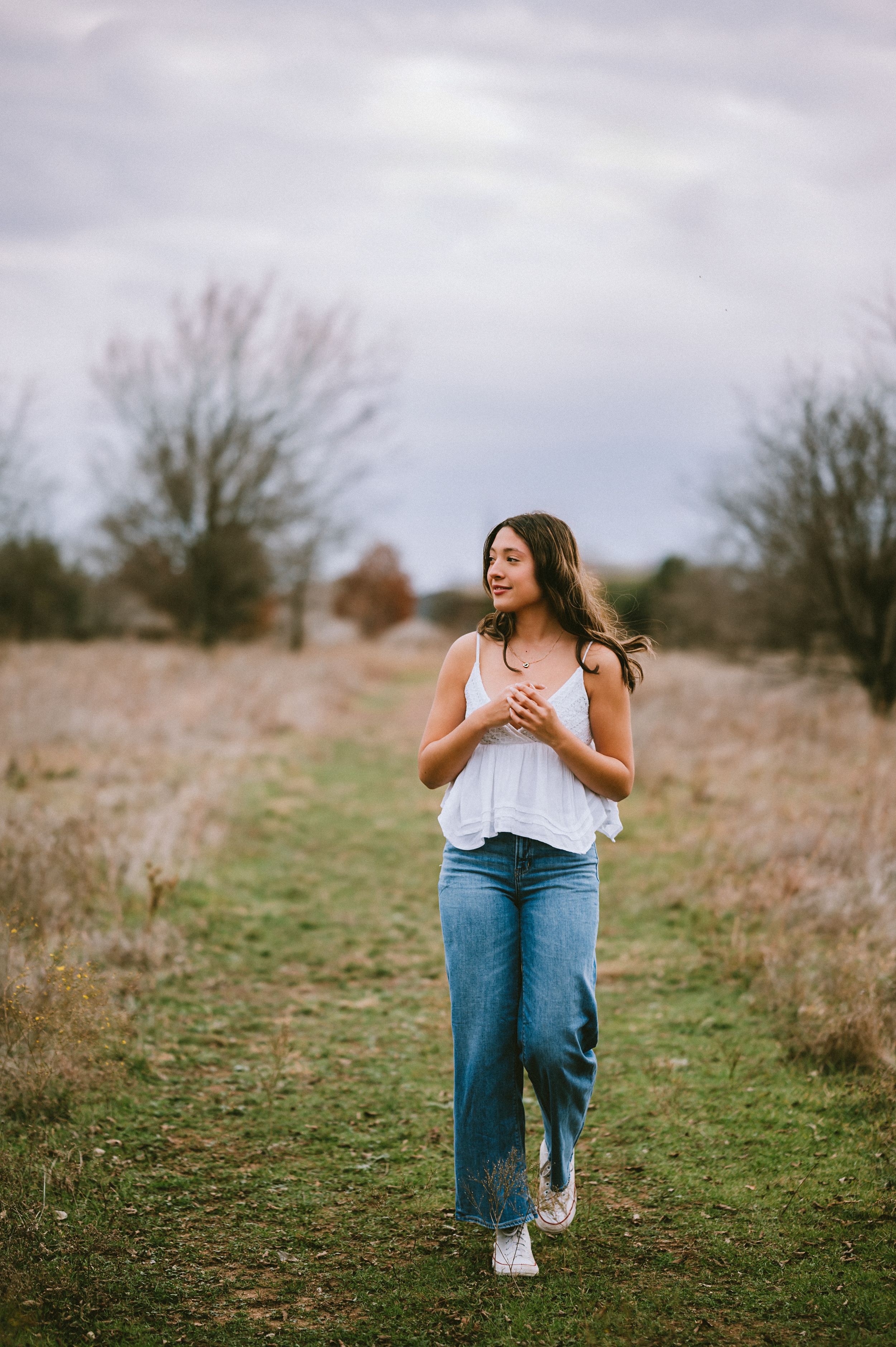 Young woman in white tank top and jeans walking along grassy rural path outdoors.