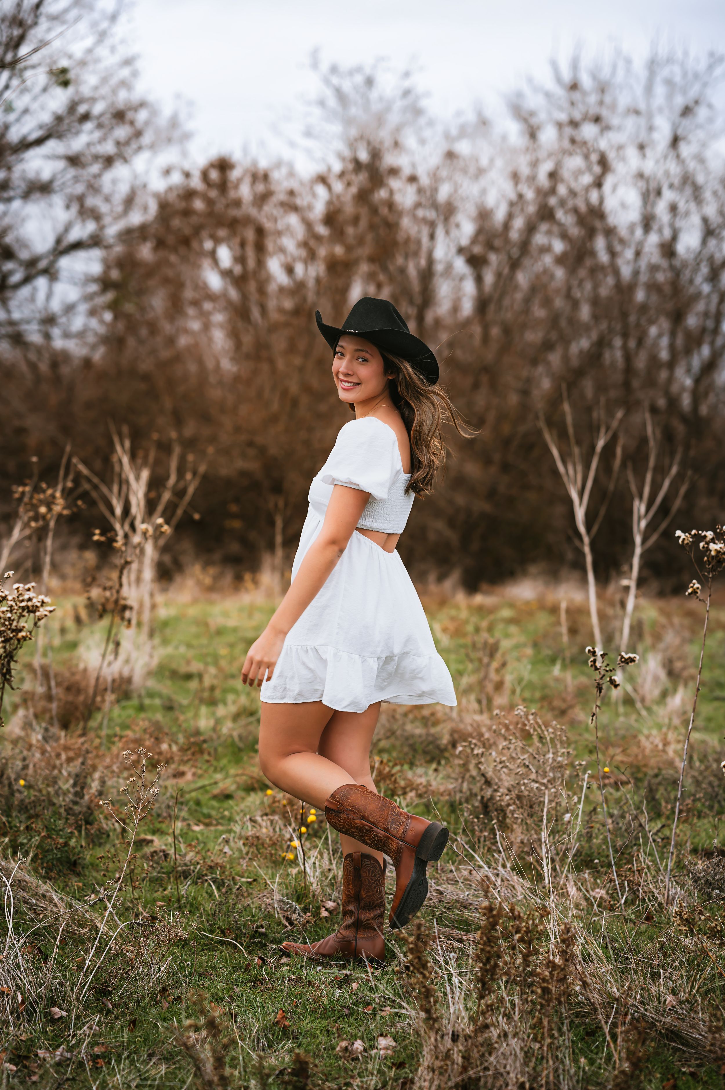 Smiling young woman in white off-shoulder dress, black cowboy hat, and brown boots in field.