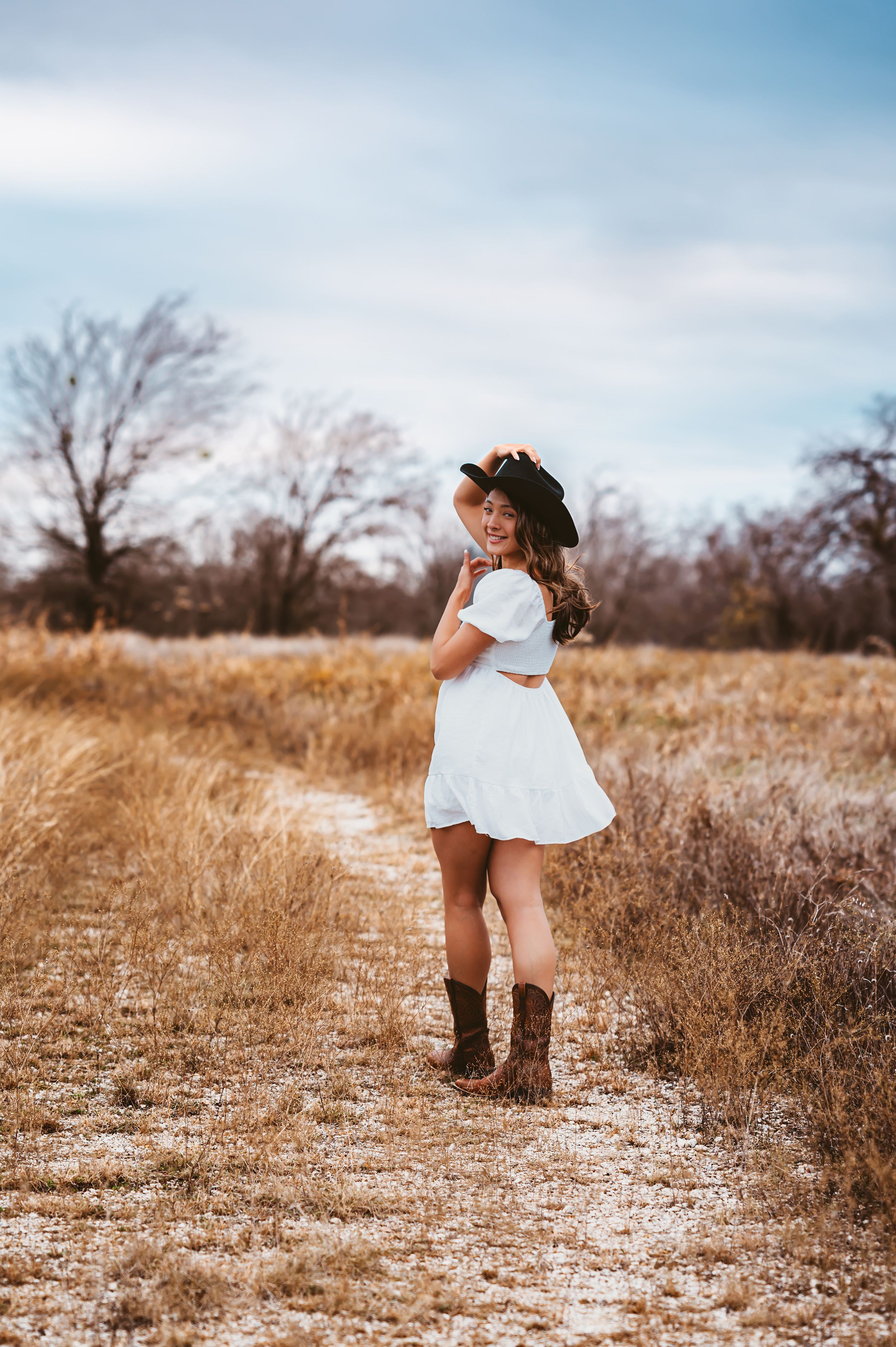 Young woman in white dress and cowboy hat with boots posing on rural dirt path.