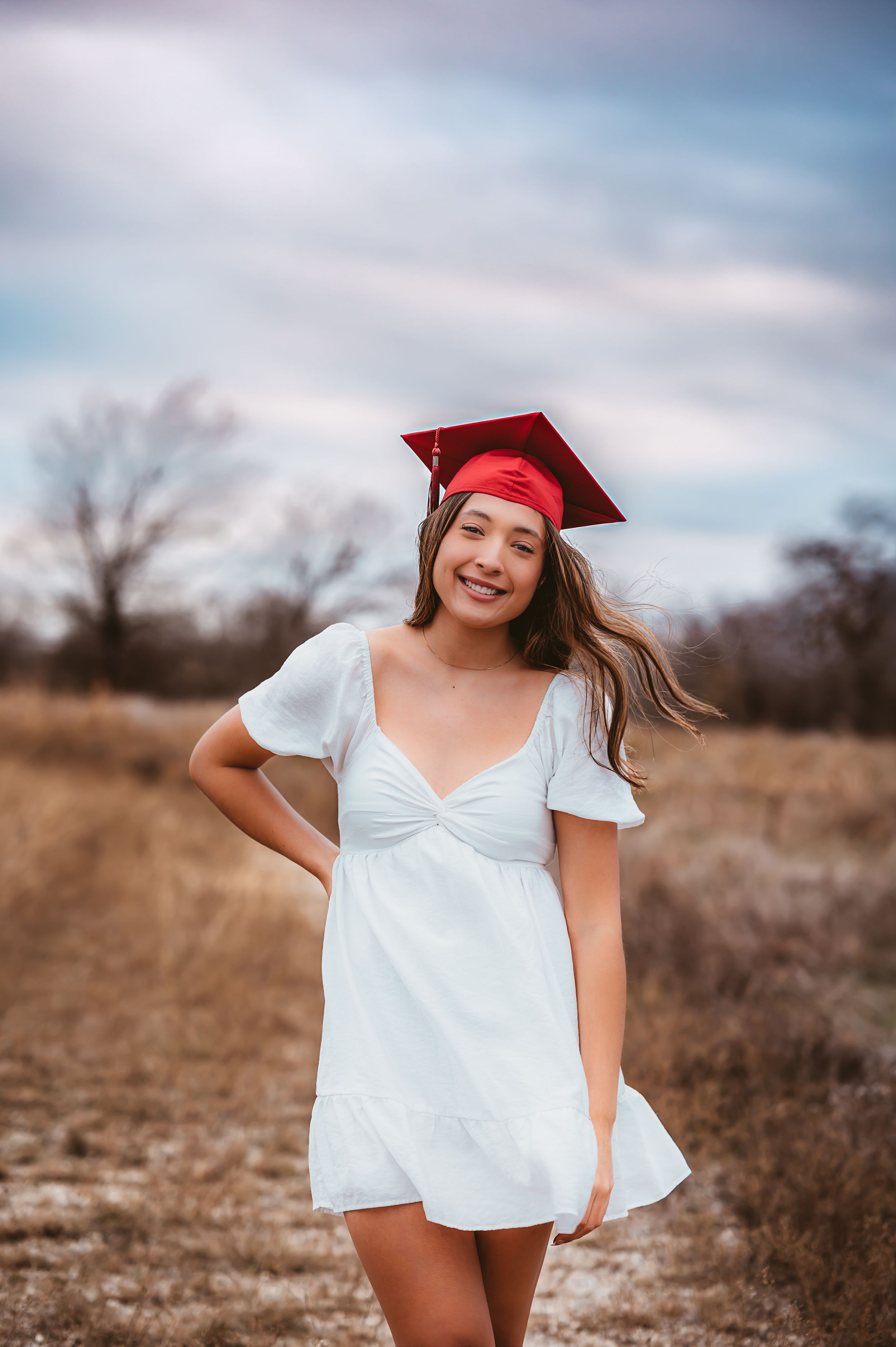 Smiling young woman in white dress and red graduation cap standing in golden field.