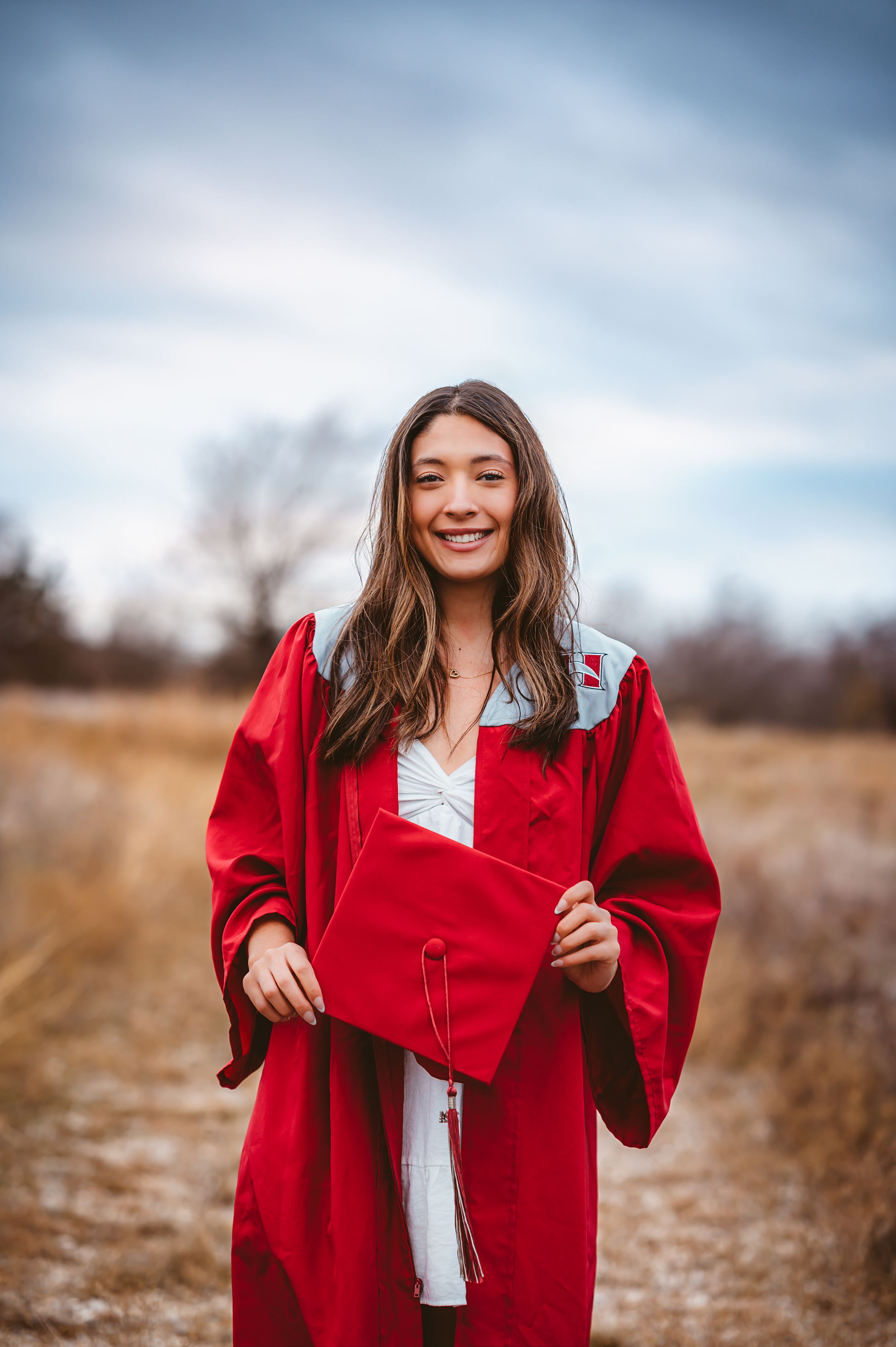Happy young woman in red graduation cap and gown holding cap in open field.