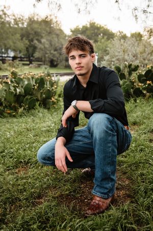 Young man in black shirt and jeans crouching in grass near cacti for a senior portrait.