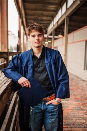 Young man in blue graduation gown holding cap outdoors on rustic wooden walkway.