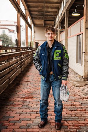 Teen boy in varsity letterman jacket holding sneakers posing on brick walkway for senior photos.