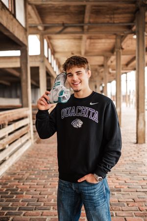 Young man in Ouachita Nike sweatshirt holding soccer ball, smiling outdoors on brick walkway.