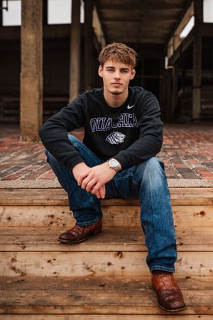 Young man in Ouachita sweatshirt and cowboy boots sitting on wooden steps with confident pose.